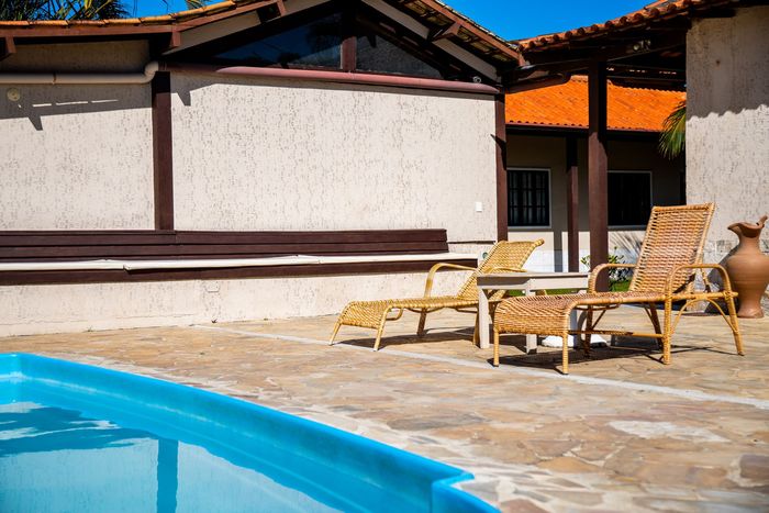 Cena à beira da piscina: duas espreguiçadeiras junto a uma piscina azul, perto de um edifício com paredes texturizadas em tons de bege e telhado de telhas vermelhas.
