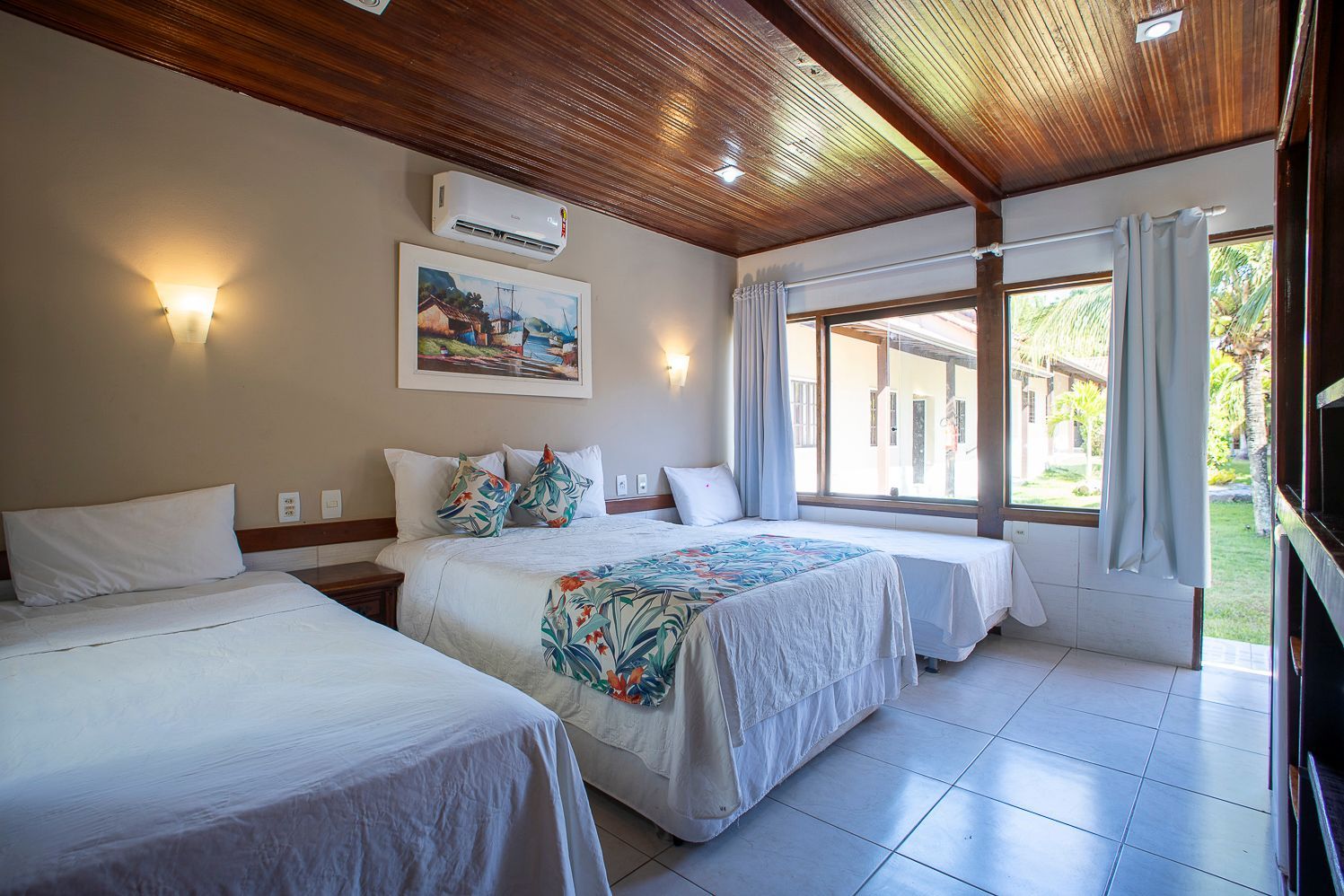 Quarto com três camas: duas de solteiro e uma de casal, paredes claras e janela com vista.