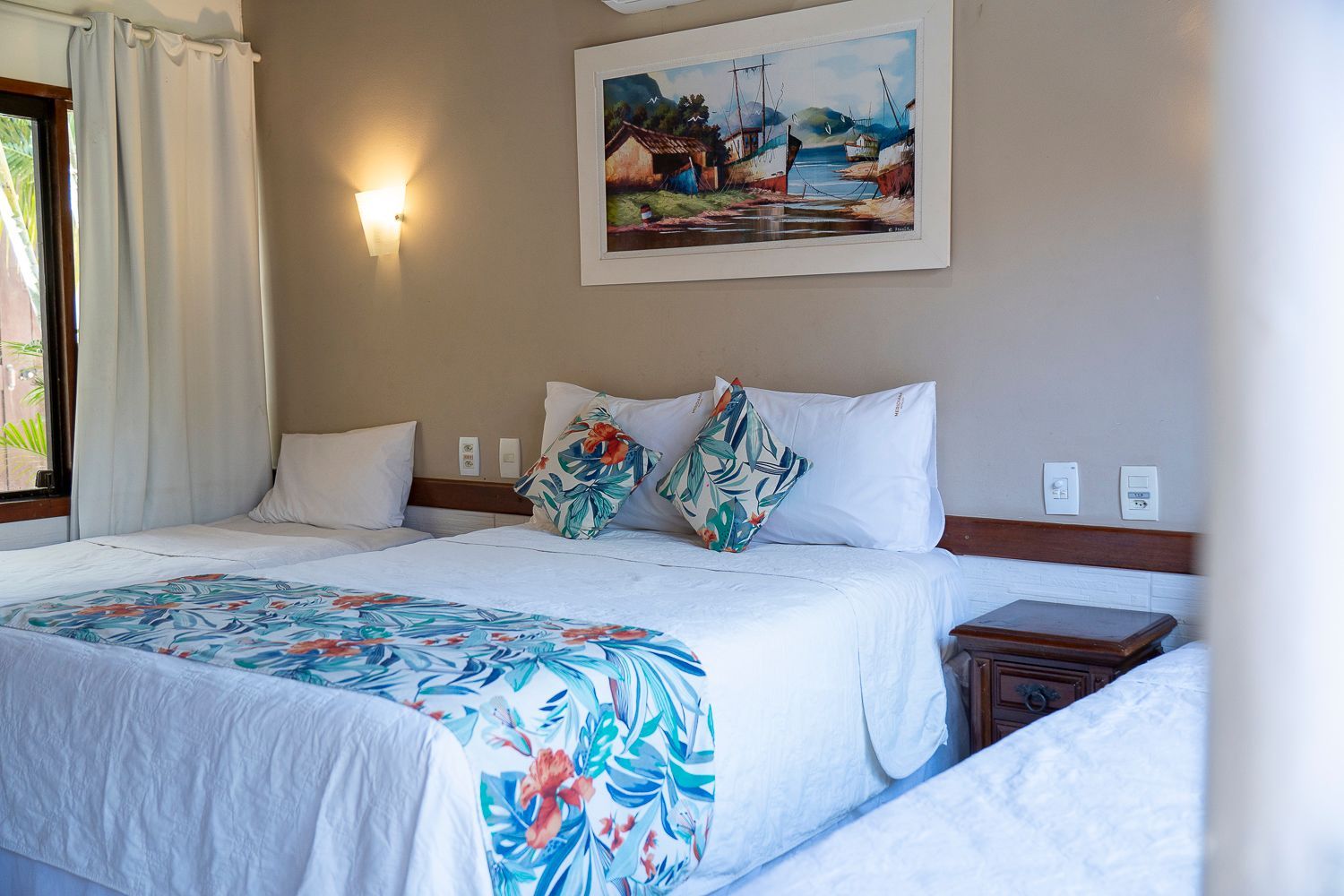 Quarto com três camas, lençóis brancos, peseira floral e decoração de parede.