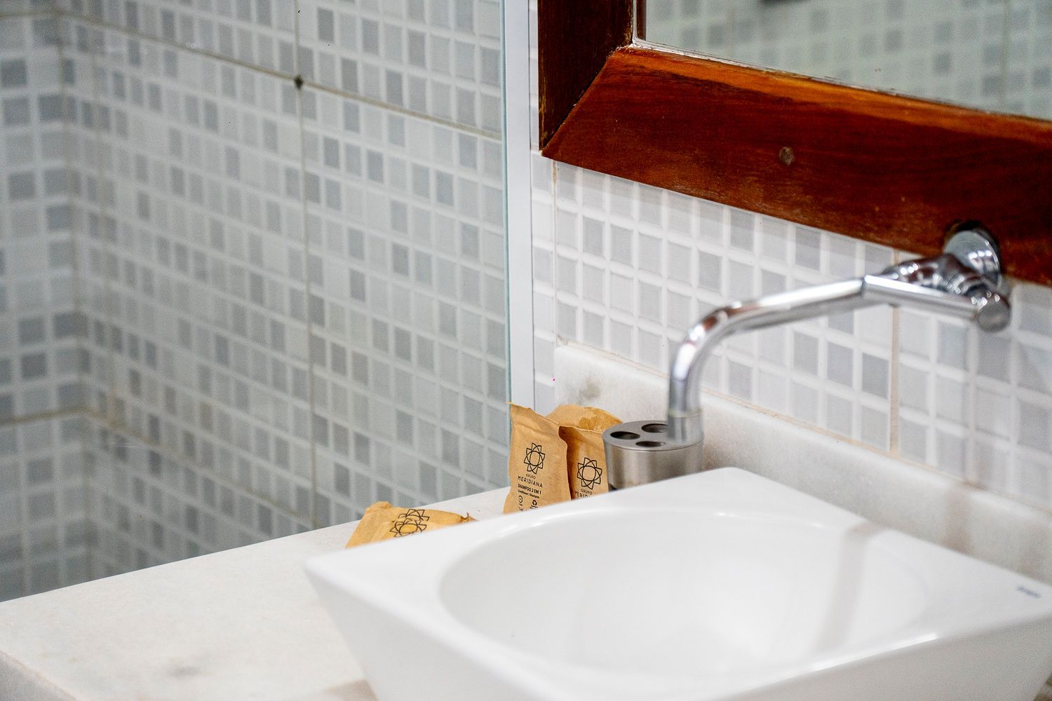 Lavatório com torneira cromada, cuba quadrada branca, parede com azulejos pequenos e espelho com moldura de madeira.