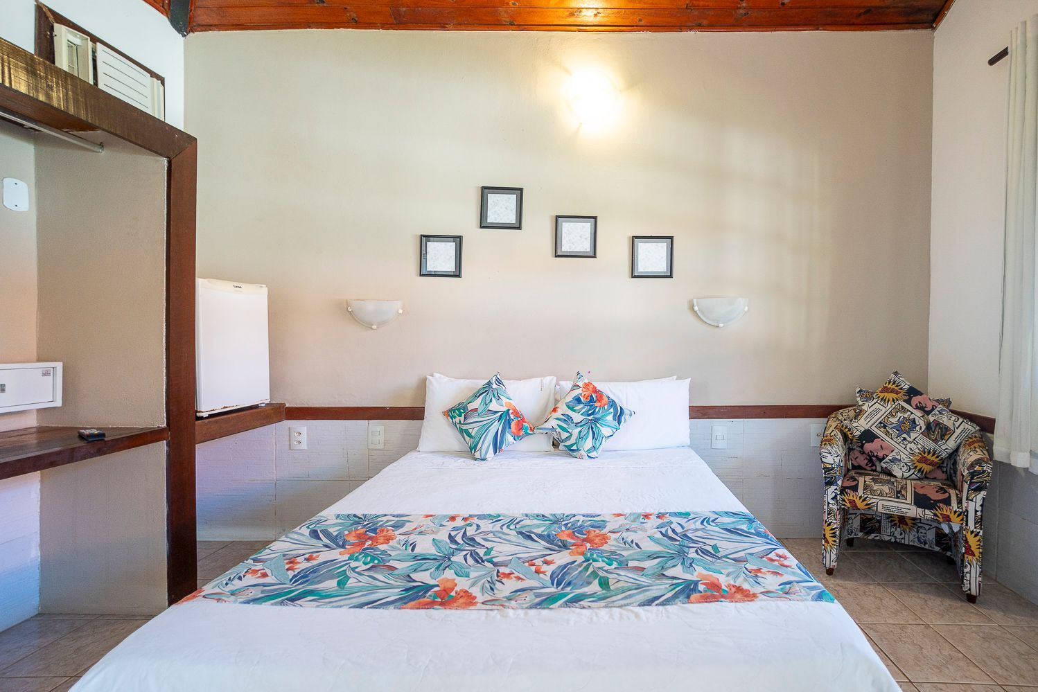 Quarto com cama de casal, peseira de cama floral, obras de arte emolduradas, poltrona estampada e paredes brancas.