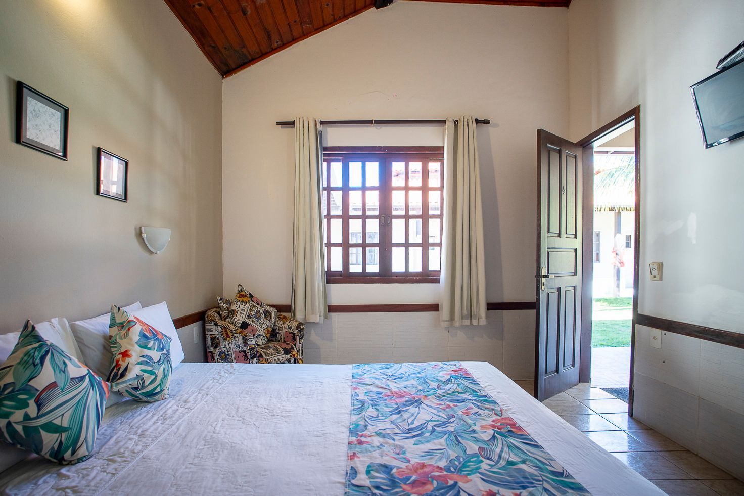 Quarto com cama, janela e porta aberta para o exterior. Paredes claras, teto de madeira.