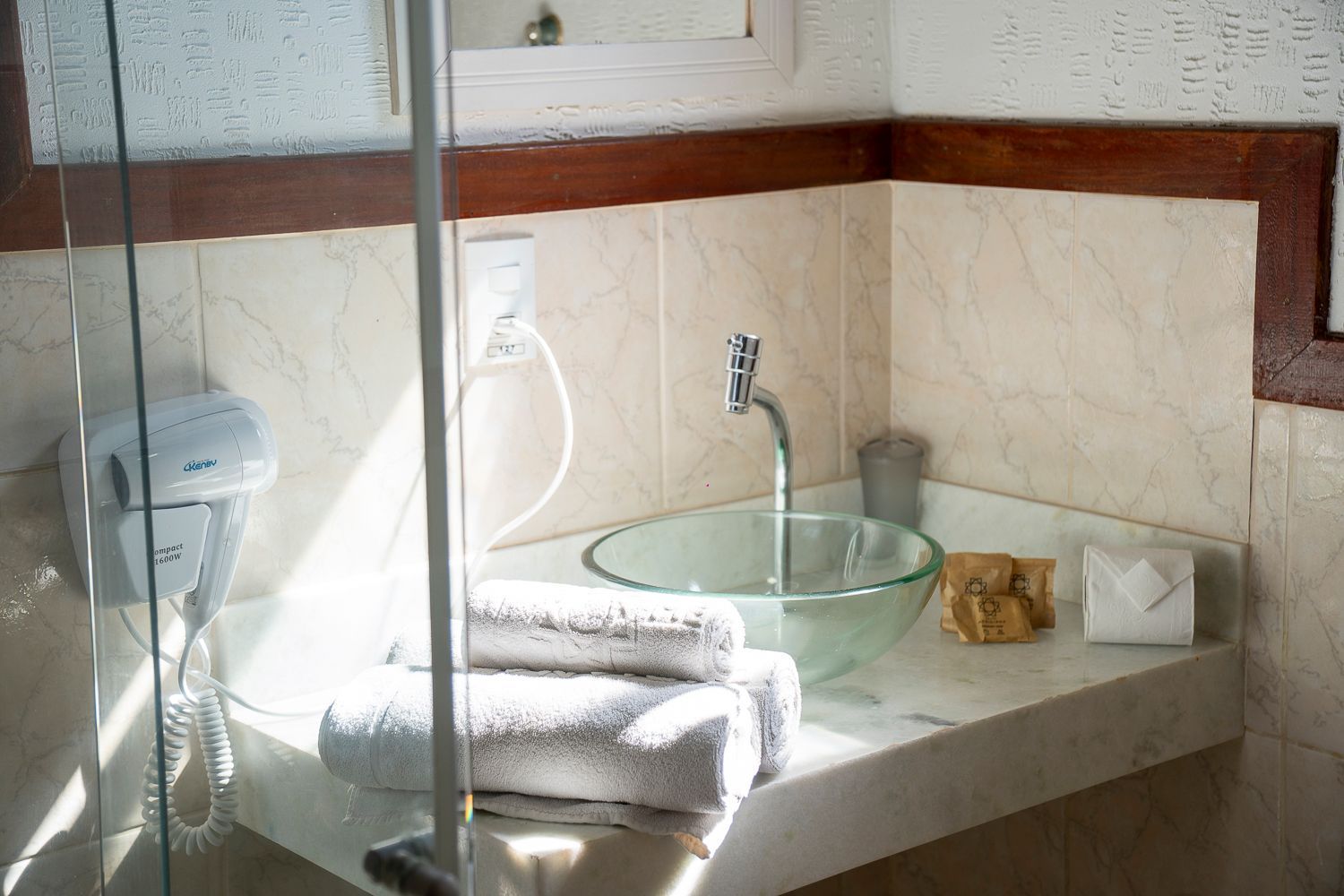 Lavatório com cuba de vidro, toalhas brancas e secador de cabelo. Azulejos bege e bancada de mármore.