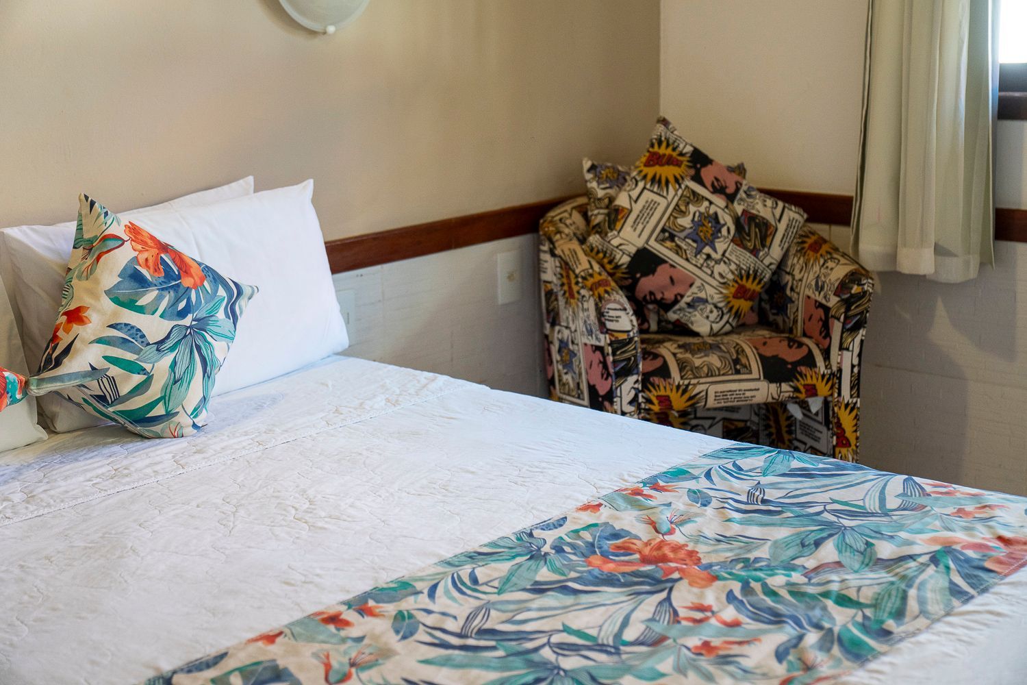 Quarto com cama branca, almofadas e manta florais e uma poltrona estampada.