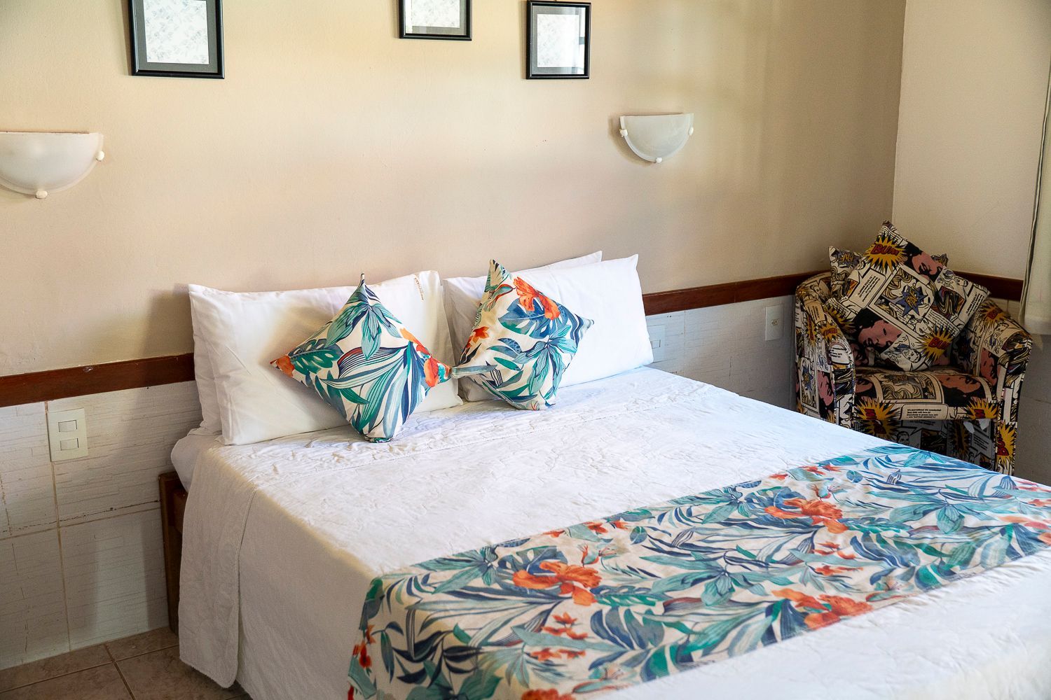 Quarto com cama, almofadas estampadas e peseira, pequeno quadro decorativo e uma poltrona estampada.
