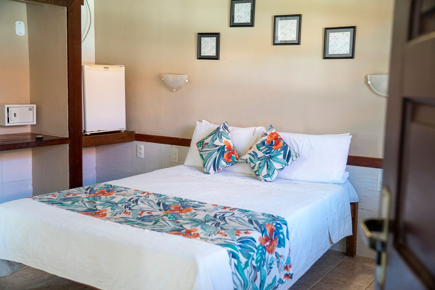 Quarto com cama, detalhes florais, obras de arte emolduradas e uma pequena geladeira.