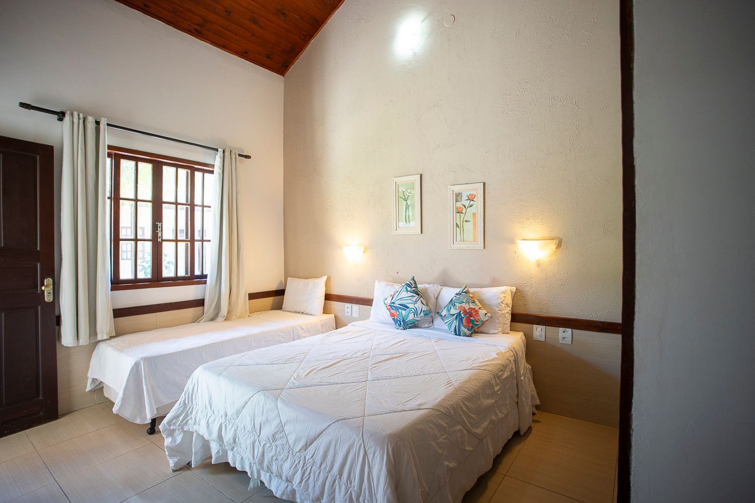 Quarto com uma cama de casal e uma de solteiro, janela e duas pequenas obras de arte.
