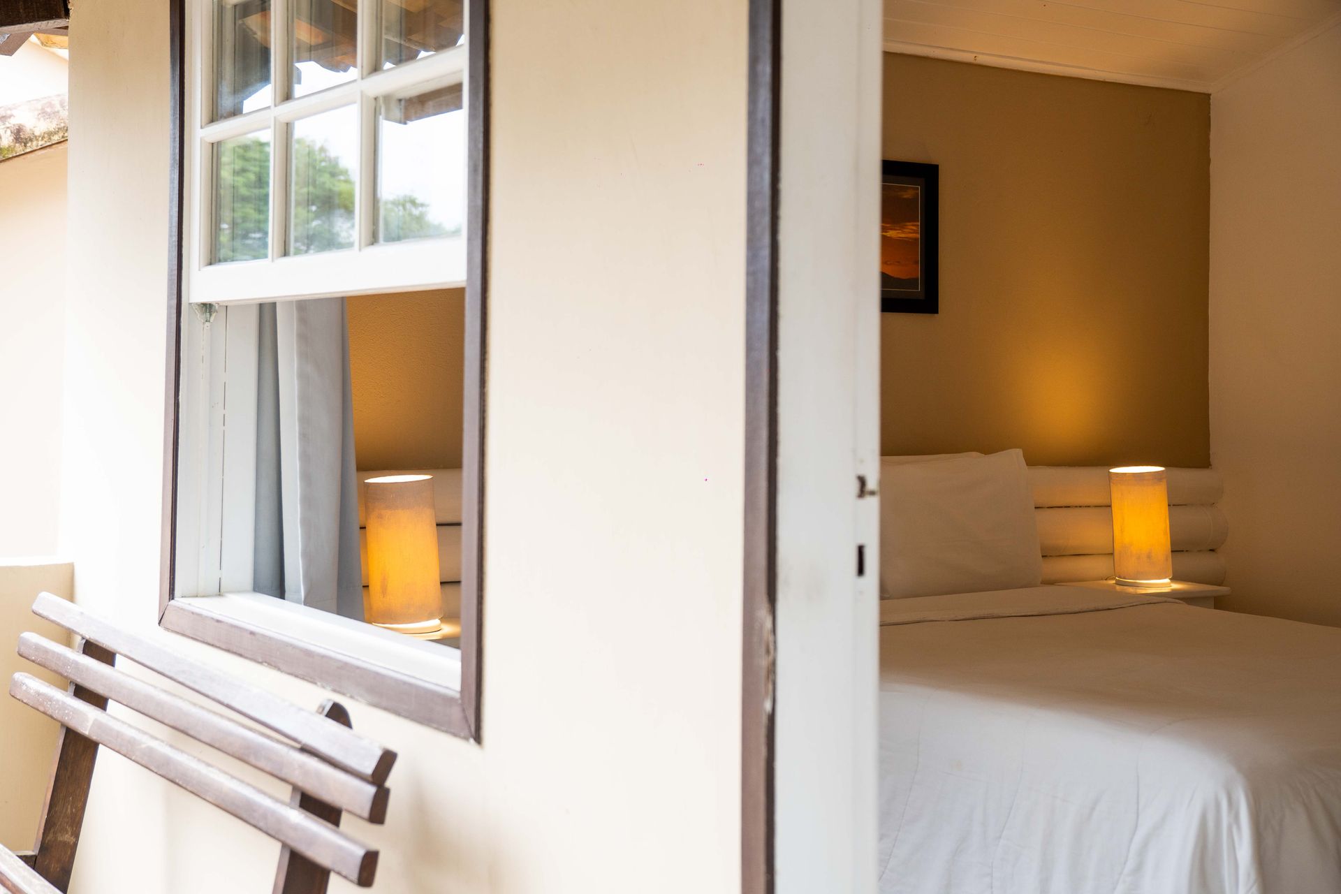 Porta de um quarto com janela; uma cama com lençóis brancos é visível, e um banco está em frente à janela.