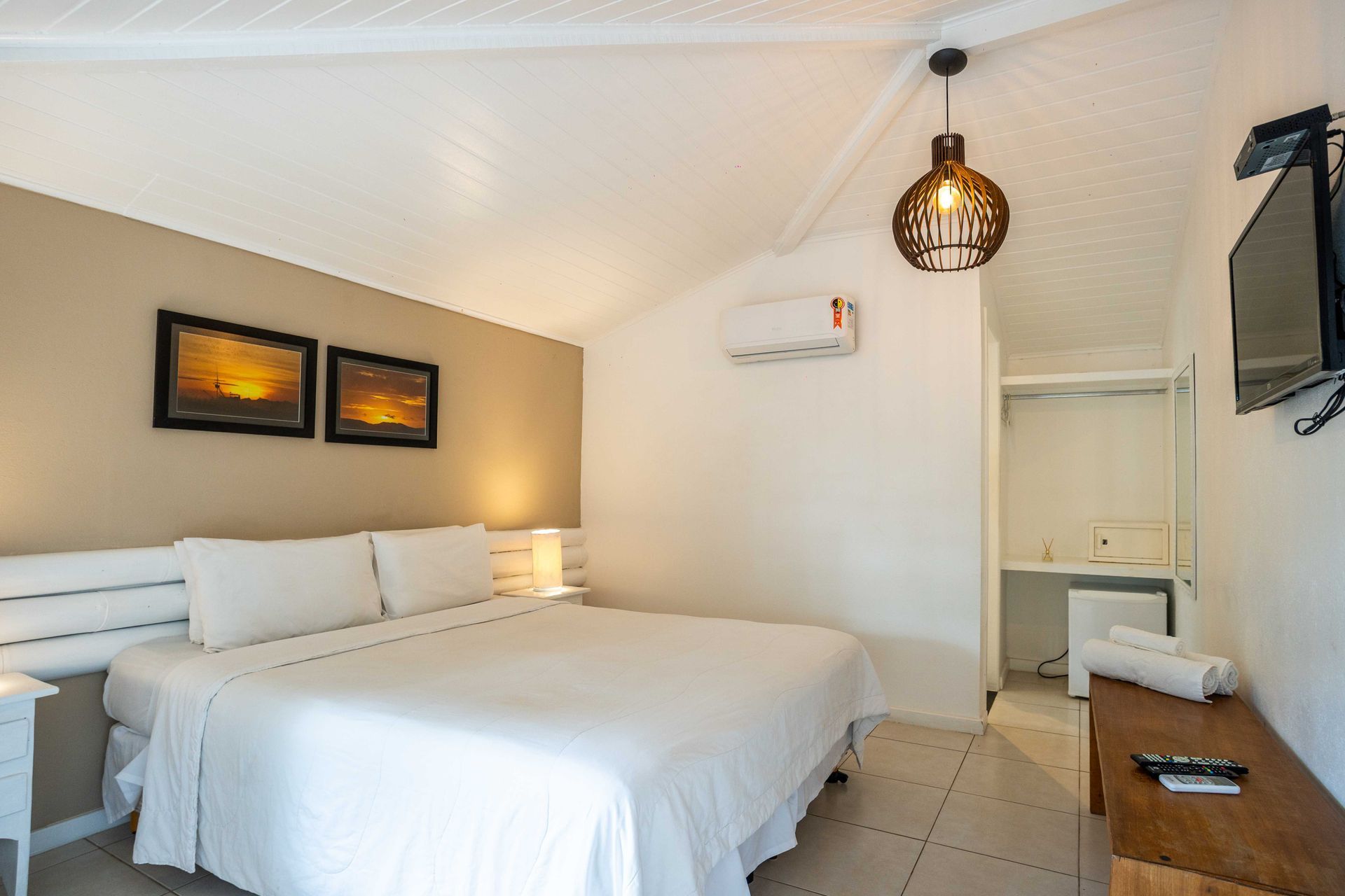Quarto de hotel com paredes brancas, cama, TV e obras de arte. Possui ar-condicionado e luminária de teto.