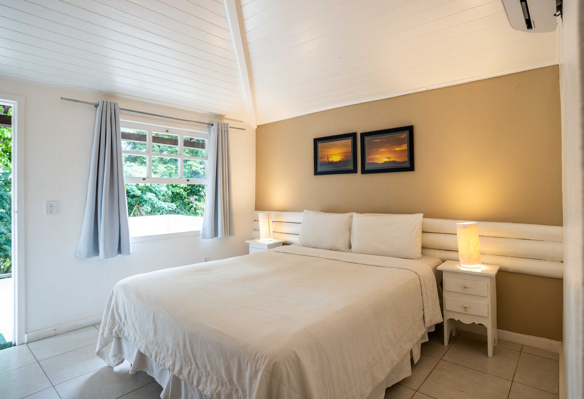 Quarto com cama branca, parede bege, janela com cortinas e obras de arte.