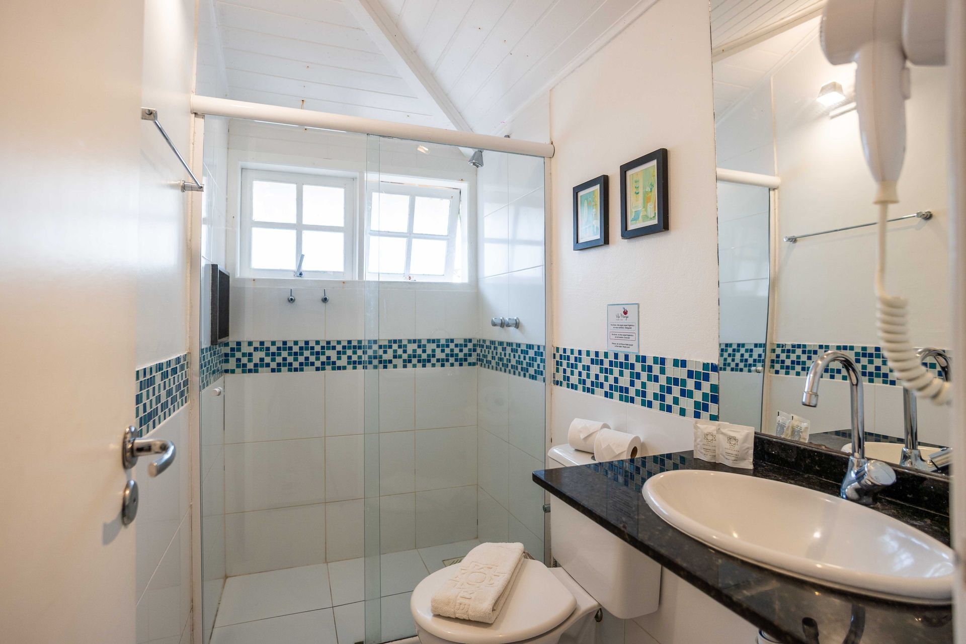 Banheiro com chuveiro, vaso sanitário e pia. Paredes brancas com detalhes em azulejo azul. Inclui janela, espelho e secador de cabelo.