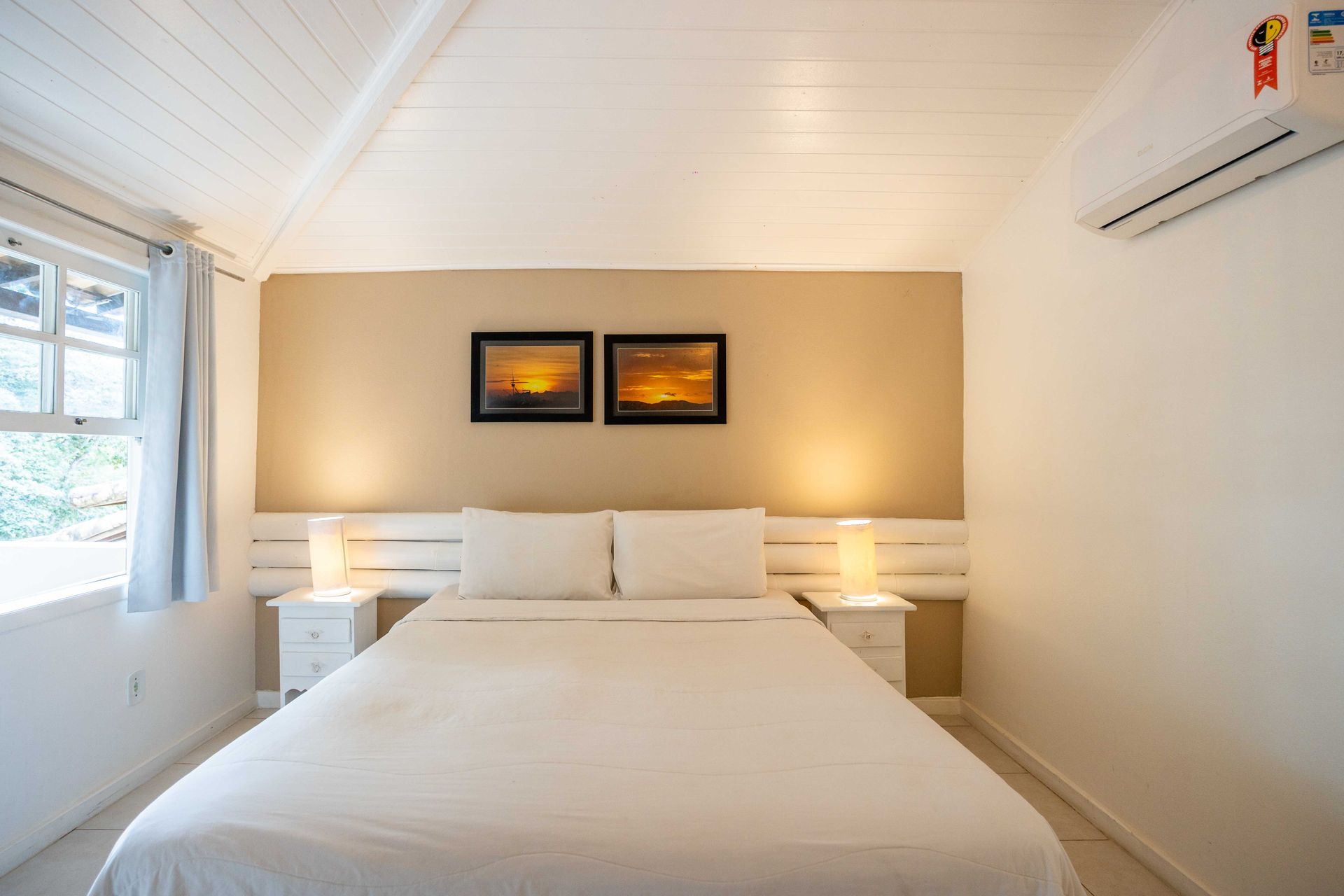 Quarto com cama branca, parede bege com obras de arte e janela com luz natural.