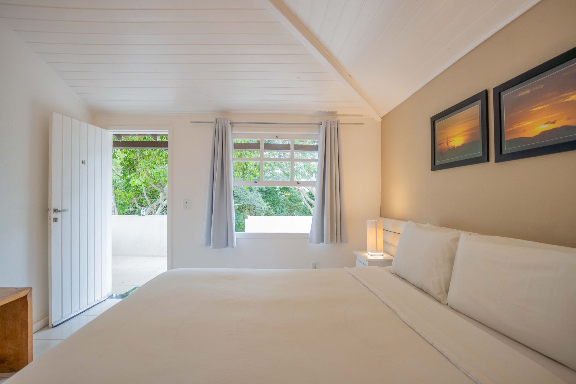 Quarto com porta e janela abertas; cama branca, paredes em tons neutros, obras de arte emolduradas.