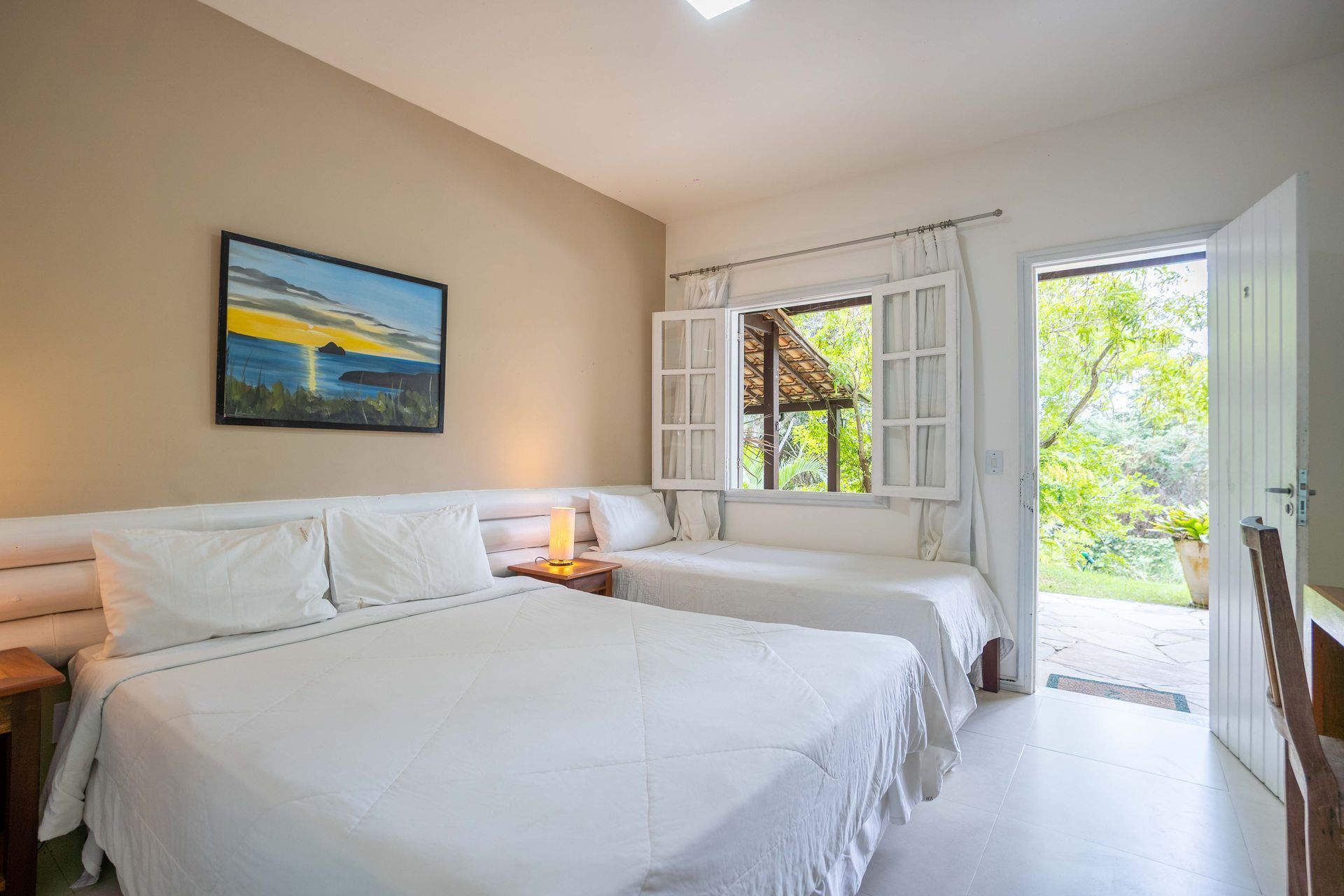 Quarto com três camas, porta aberta para a área verde e janela com vista para uma varanda.