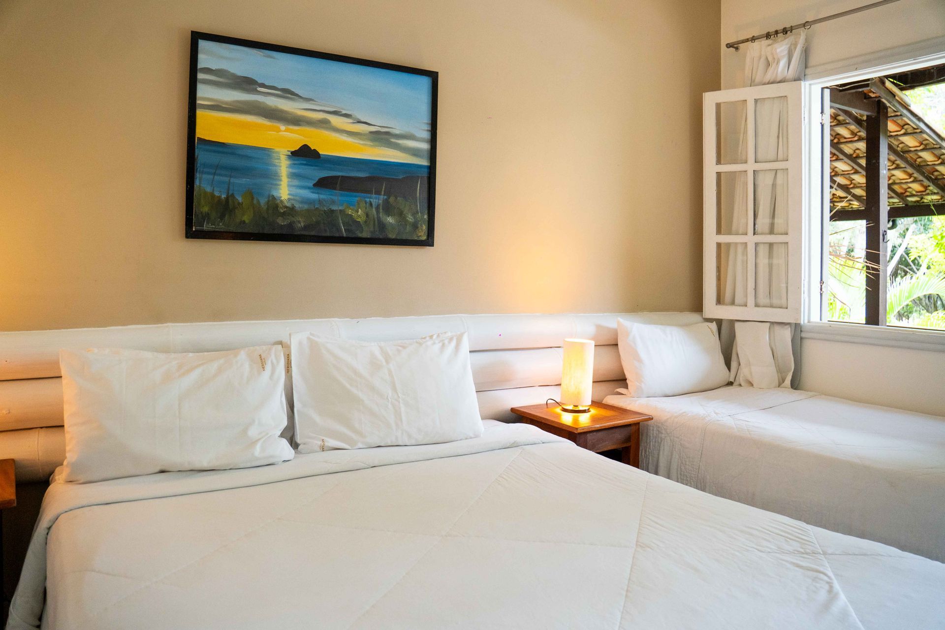 Quarto com duas camas, lençóis brancos, pintura de uma paisagem marítima e luz natural proveniente de uma janela.