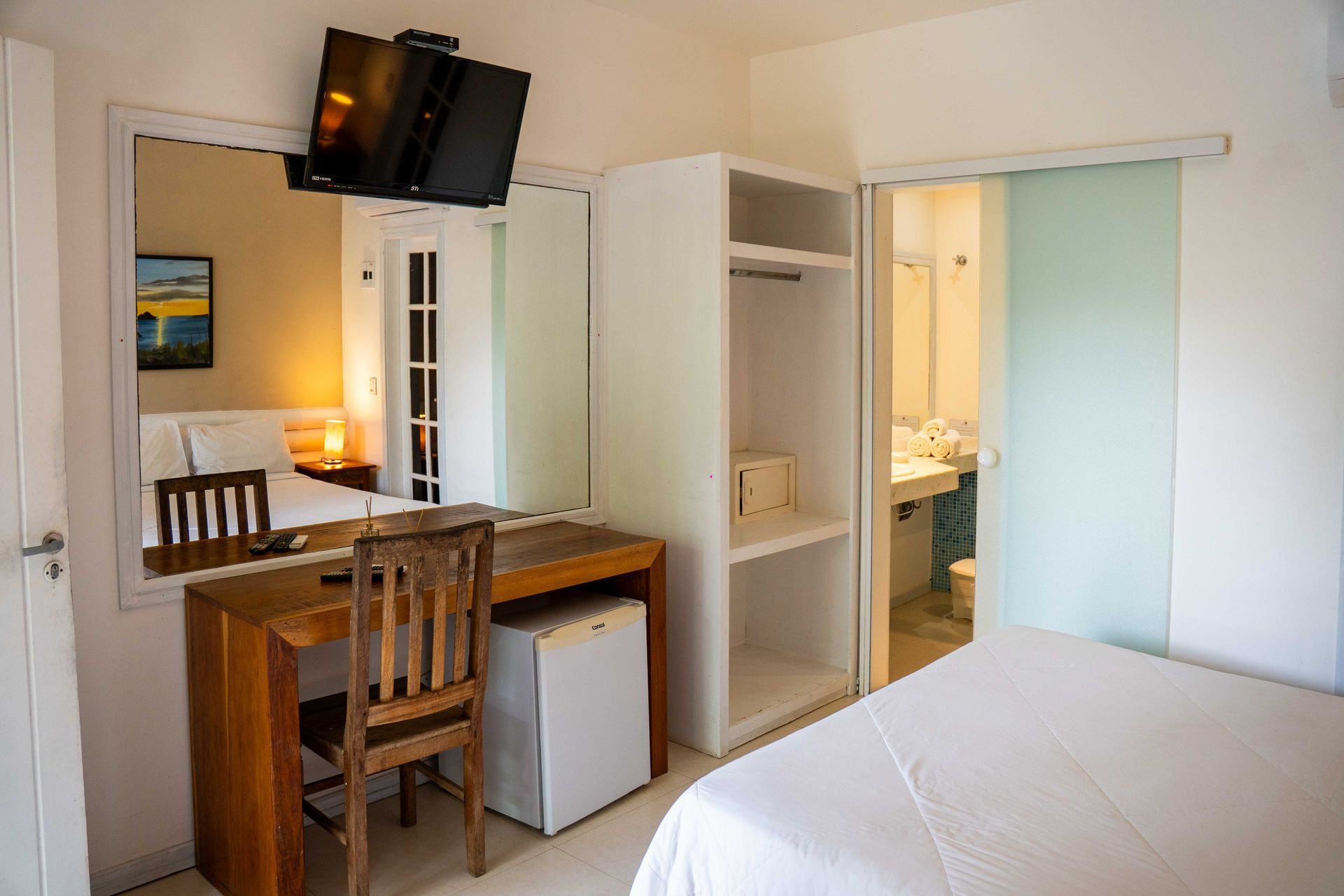 Quarto de hotel com cama, escrivaninha, espelho, armário e banheiro visíveis através de uma porta de correr.