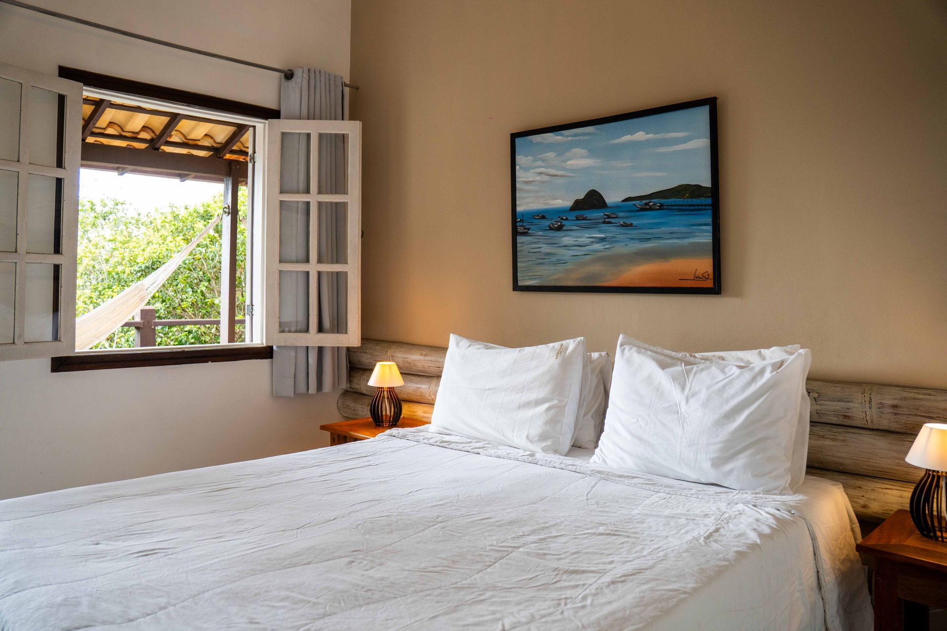 Quarto com roupa de cama branca, janela aberta, pintura de paisagem acima da cama e abajures.