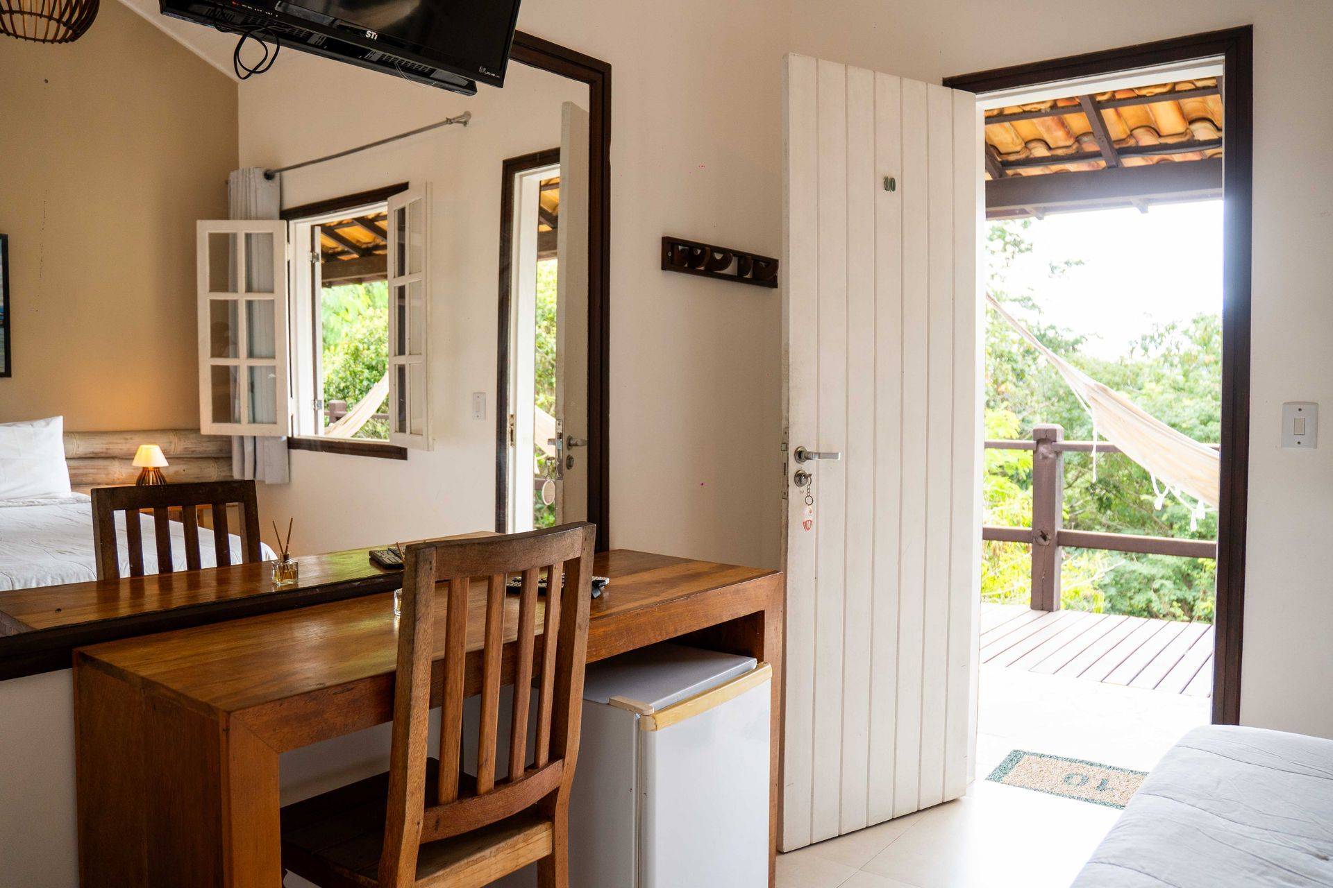 Quarto aconchegante com escrivaninha, porta aberta para varanda com rede, janela, porta branca e móveis de madeira.
