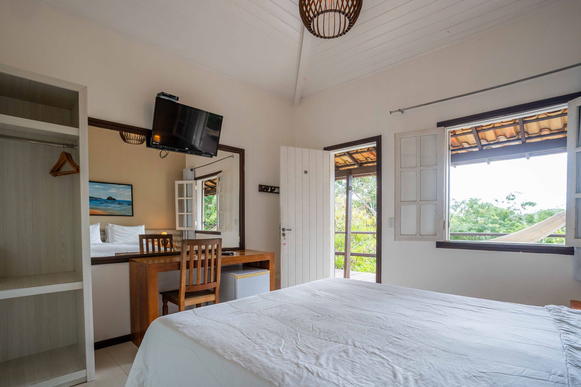 Quarto com cama, escrivaninha, janelas abertas e armário; paredes brancas, luz natural.