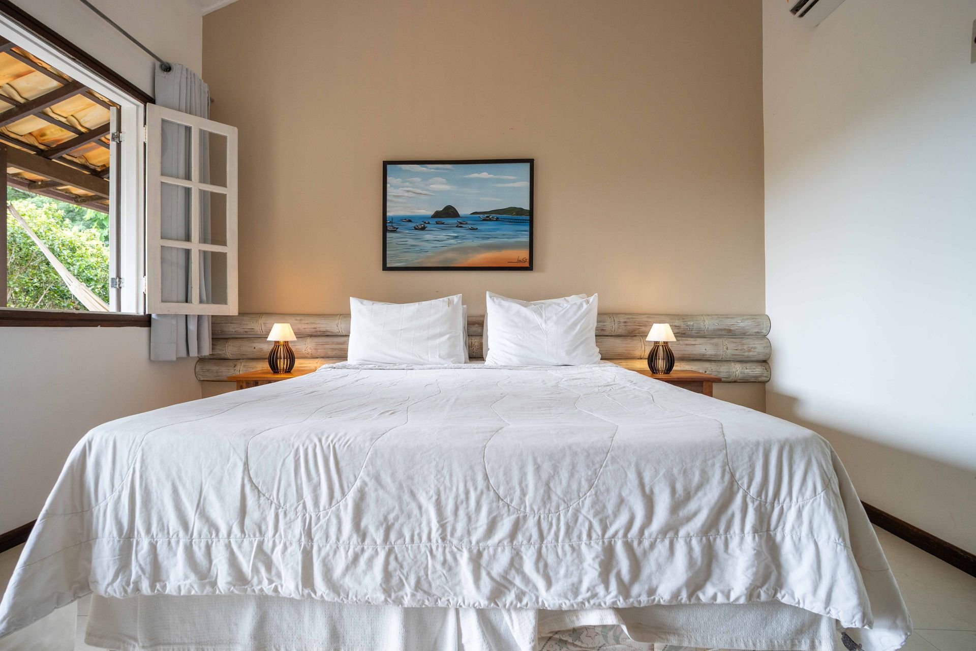 Quarto com cama king-size, lençóis brancos, dois abajures, um quadro e uma janela com veneziana aberta.