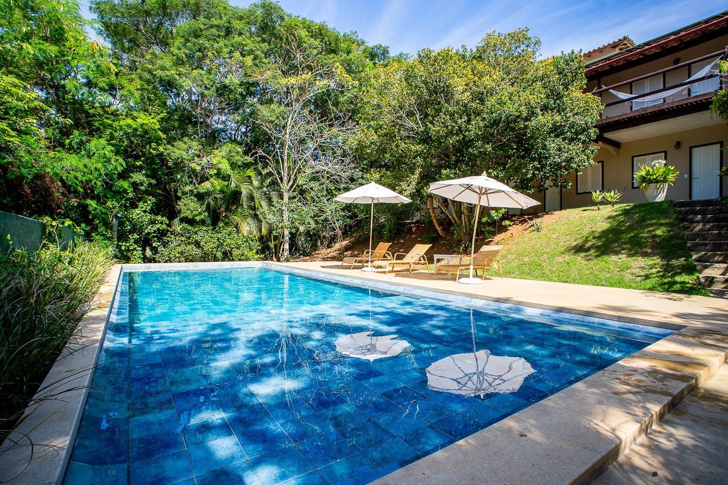 Piscina com azulejos azuis rodeada de vegetação e um edifício. Dispõe de dois guarda-sóis e espreguiçadeiras.