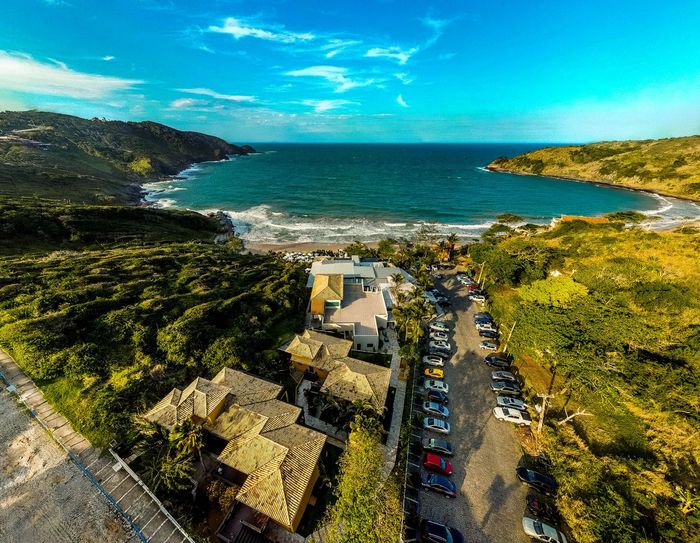 Vista aérea de uma praia com casas, carros e vegetação exuberante sob um céu azul claro.