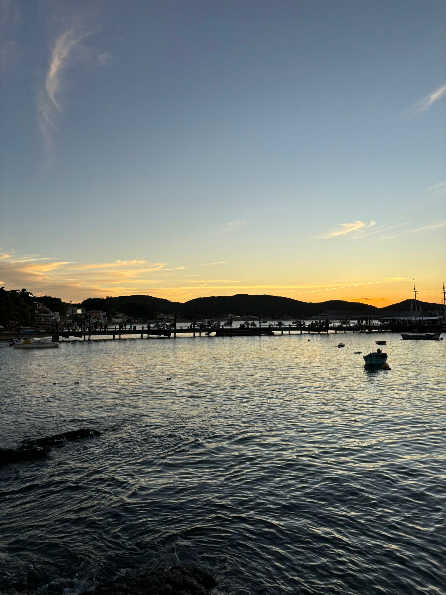 Pôr do sol sobre o porto, silhueta de docas e edifícios. Água calma, barcos. Céu laranja e azul.