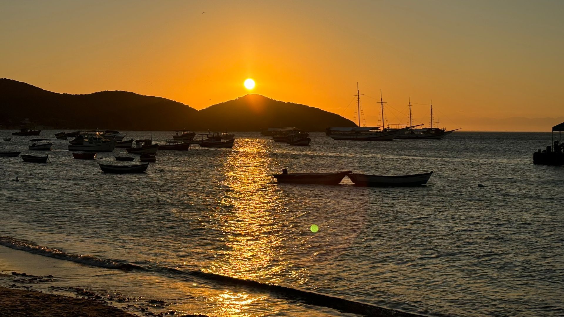 Pôr do sol sobre o oceano calmo com barcos. Sol alaranjado sobre colinas recortadas e barcos balançando na água.