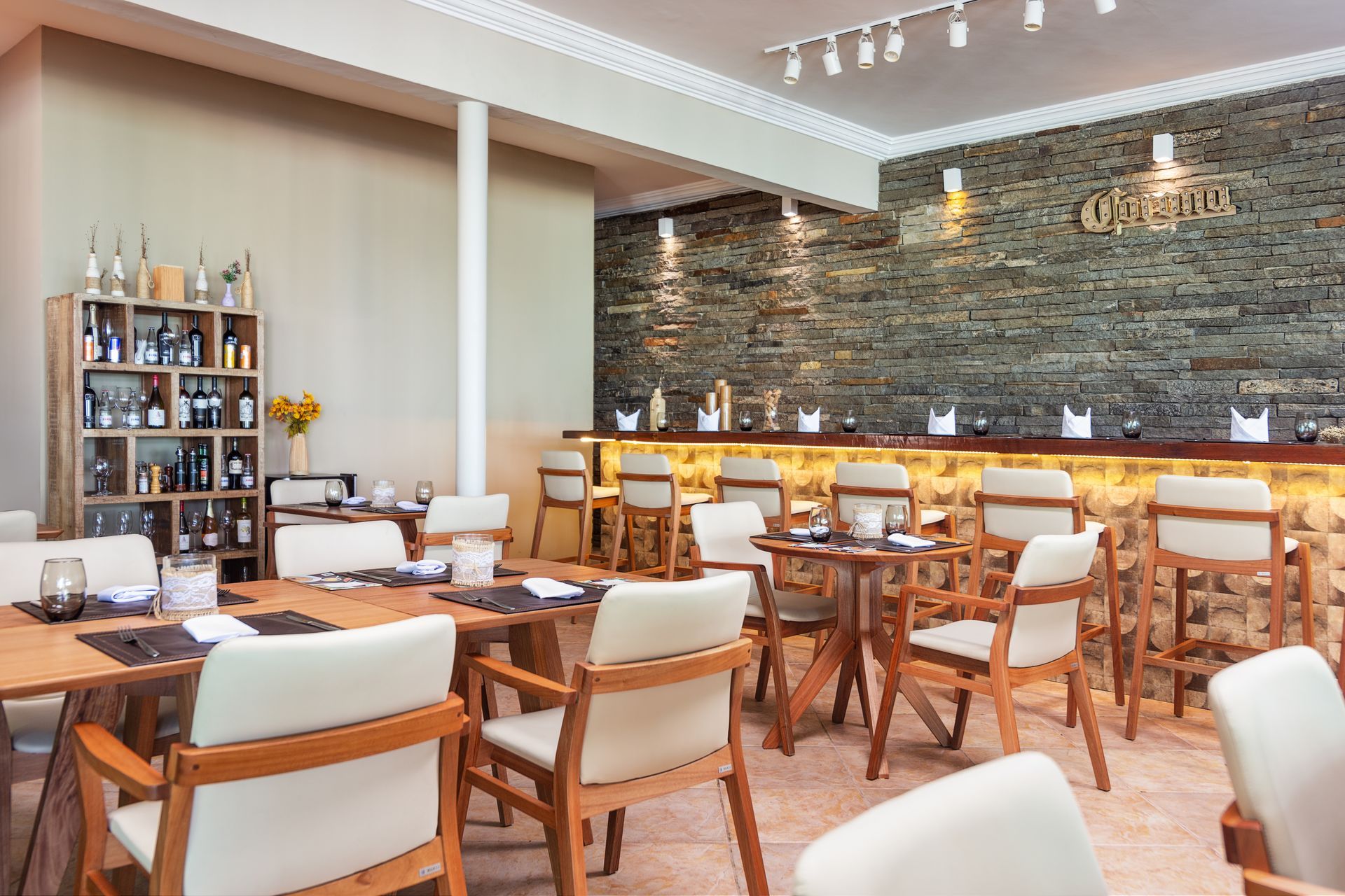Interior de restaurante com mesas de madeira, bar, parede de pedra e cadeiras cor creme.