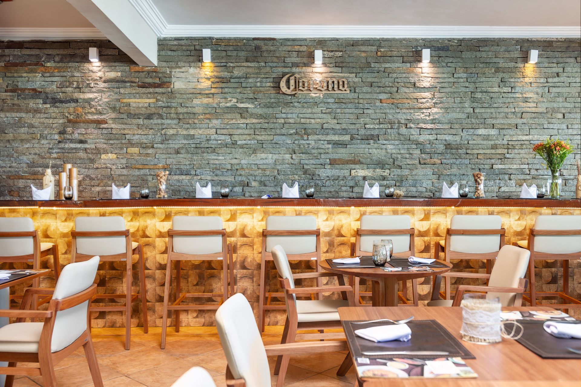 Interior de restaurante com bar e mesas. Parede de pedra atrás do bar; mesas e cadeiras de madeira.