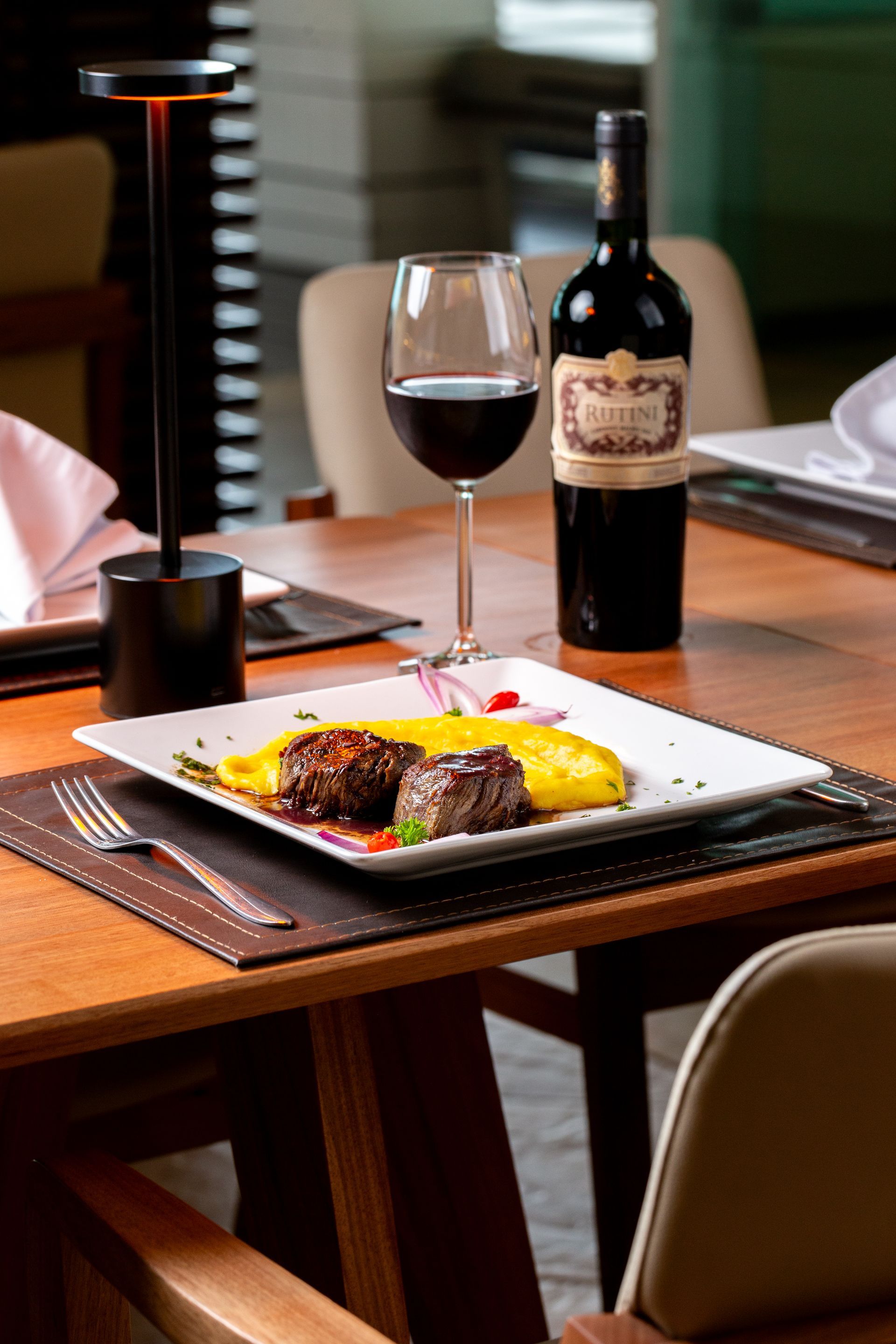 Mesa posta com bife, taça de vinho e garrafa. Mesa de madeira, cadeiras em tons neutros e um pequeno abajur.