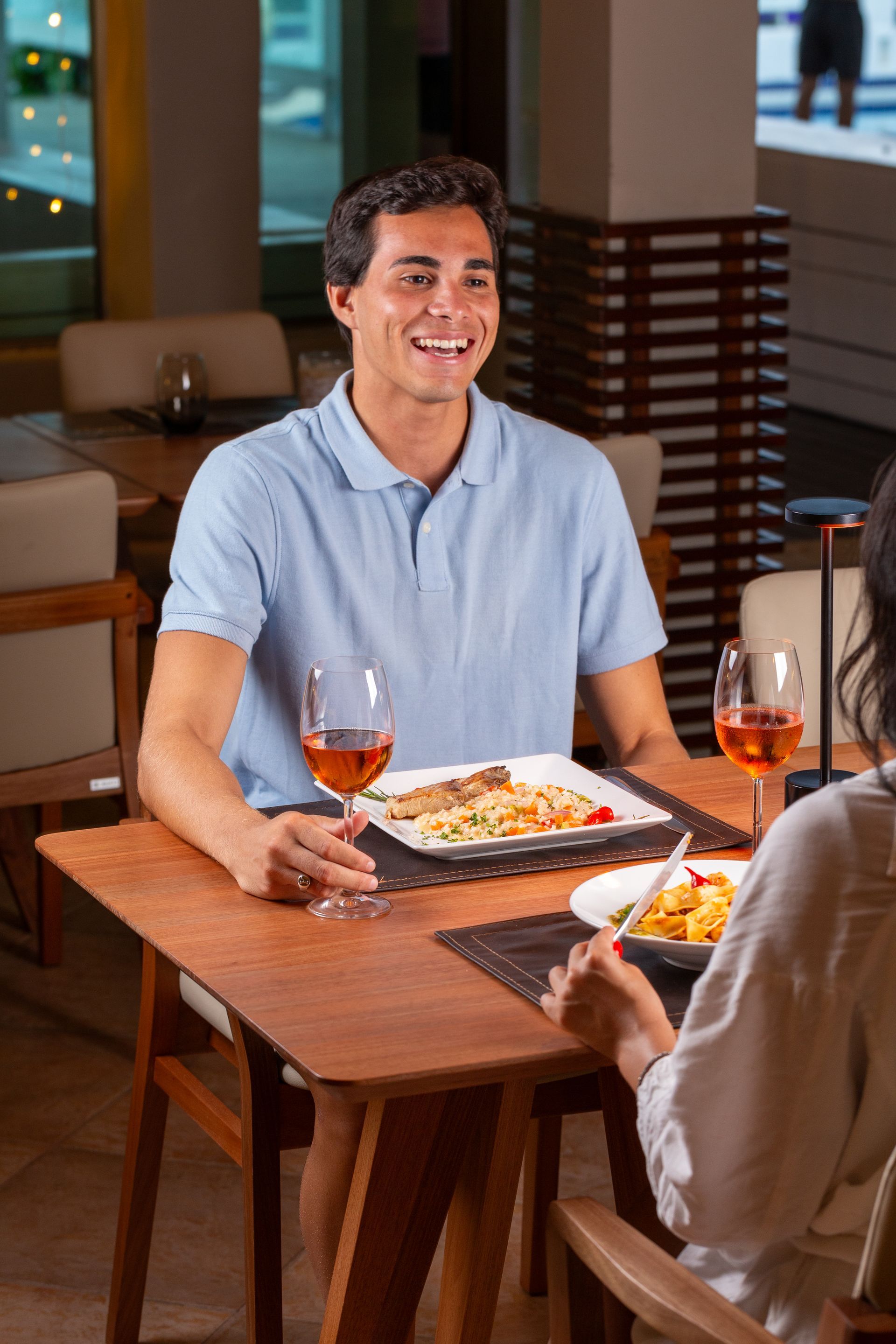 Um homem sorri enquanto janta com outra pessoa, segurando uma taça de vinho. Mesa com comida e bebidas.