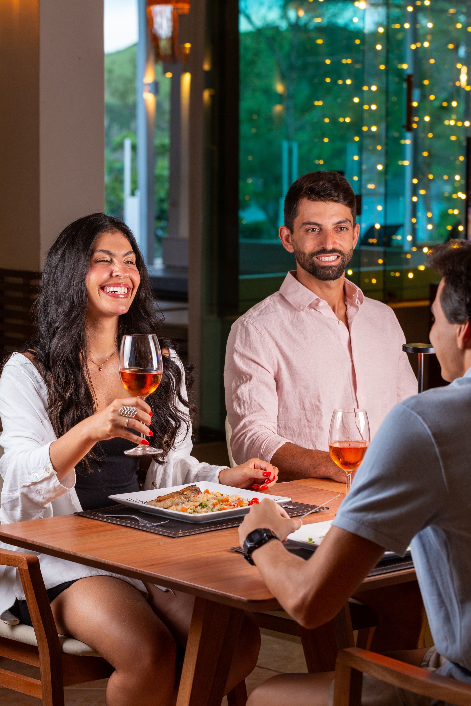 Uma mulher e um homem sorrindo, brindando com suas taças em uma mesa com comida, em um restaurante.