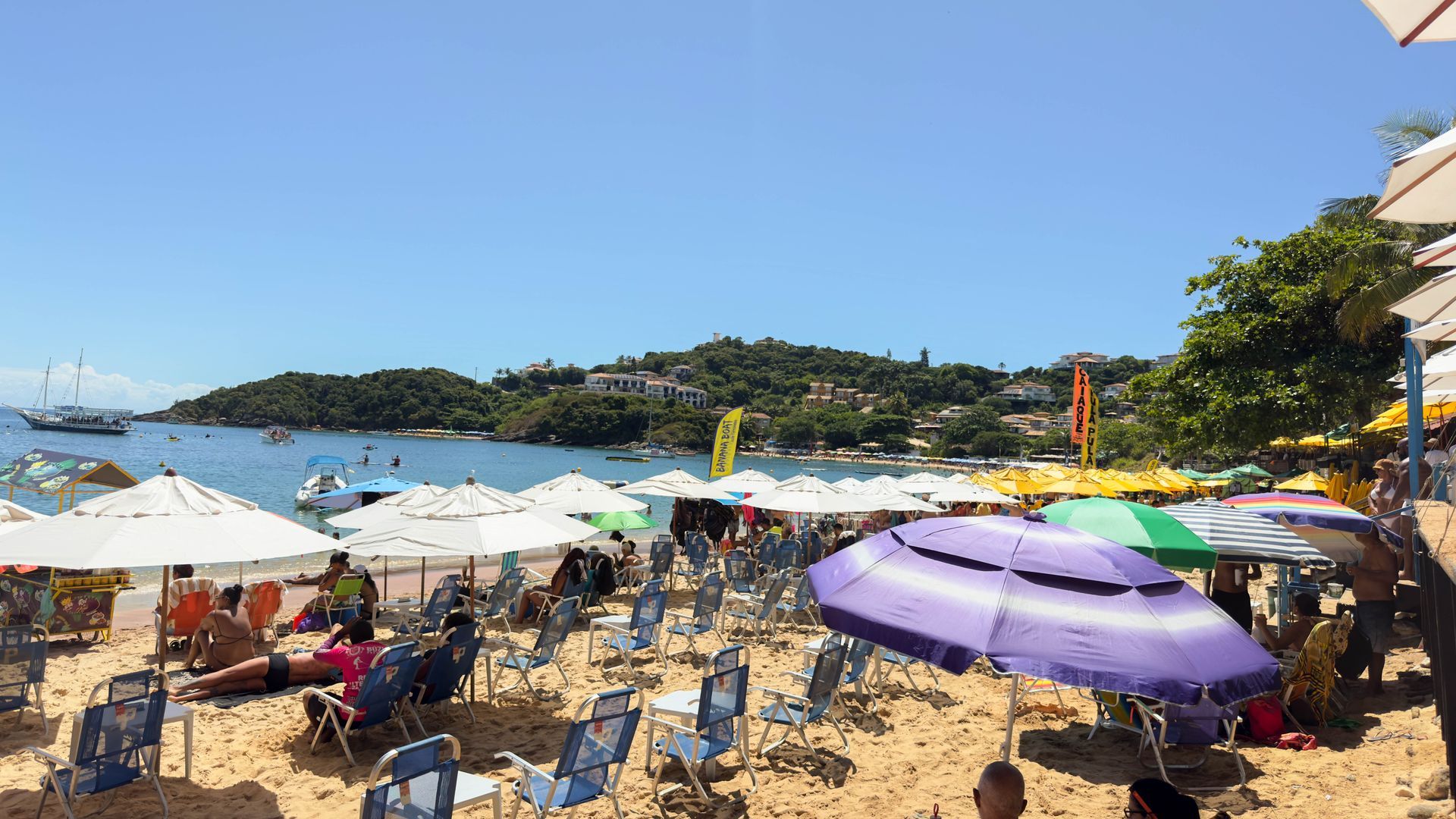Cena de praia com muitos guarda-sóis, cadeiras e pessoas; uma encosta ao fundo, barcos na água.