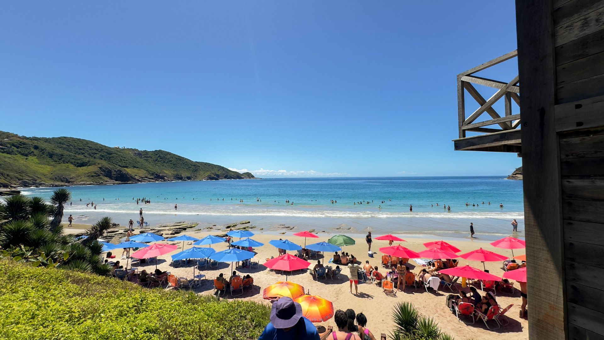 Cena de praia com guarda-sóis coloridos, pessoas e água azul cristalina sob um céu claro.