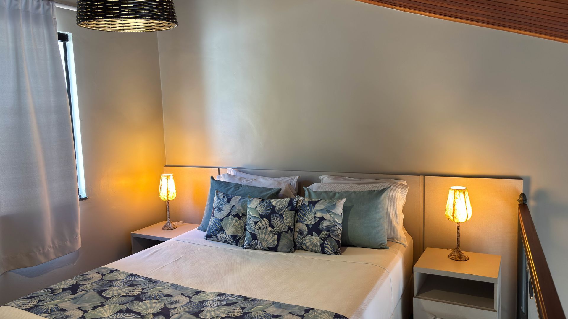 Quarto com cama, duas mesas de cabeceira, abajures, travesseiros e janela com cortinas brancas.