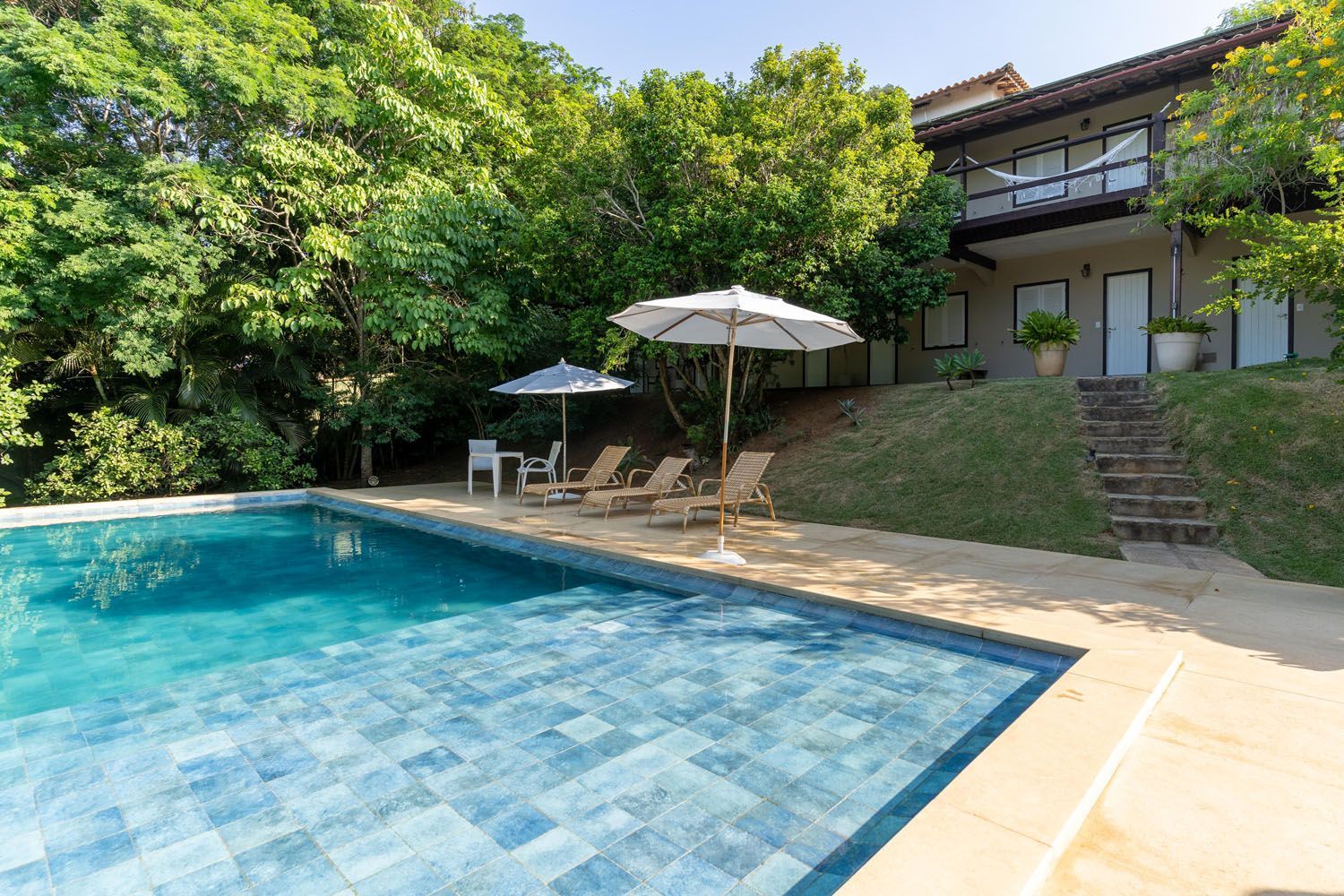 Piscina com espreguiçadeiras e guarda-sóis perto de uma casa de dois andares rodeada por árvores.