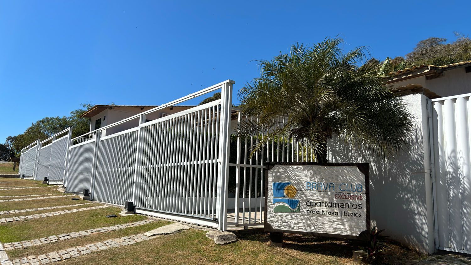 Propriedade cercada com portão e placa. A cerca é branca com um desenho geométrico e está parcialmente aberta. Céu azul ao fundo.