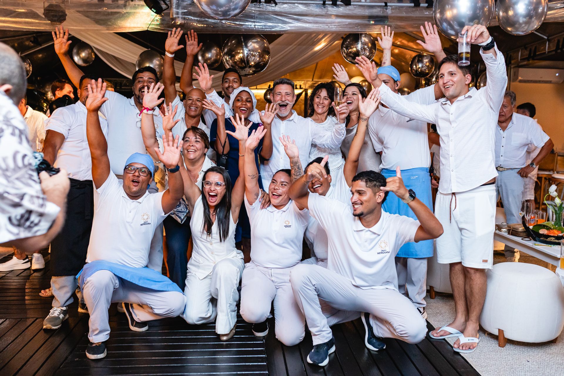 Group of people in white clothing, arms raised, celebrating indoors with silver balloons.