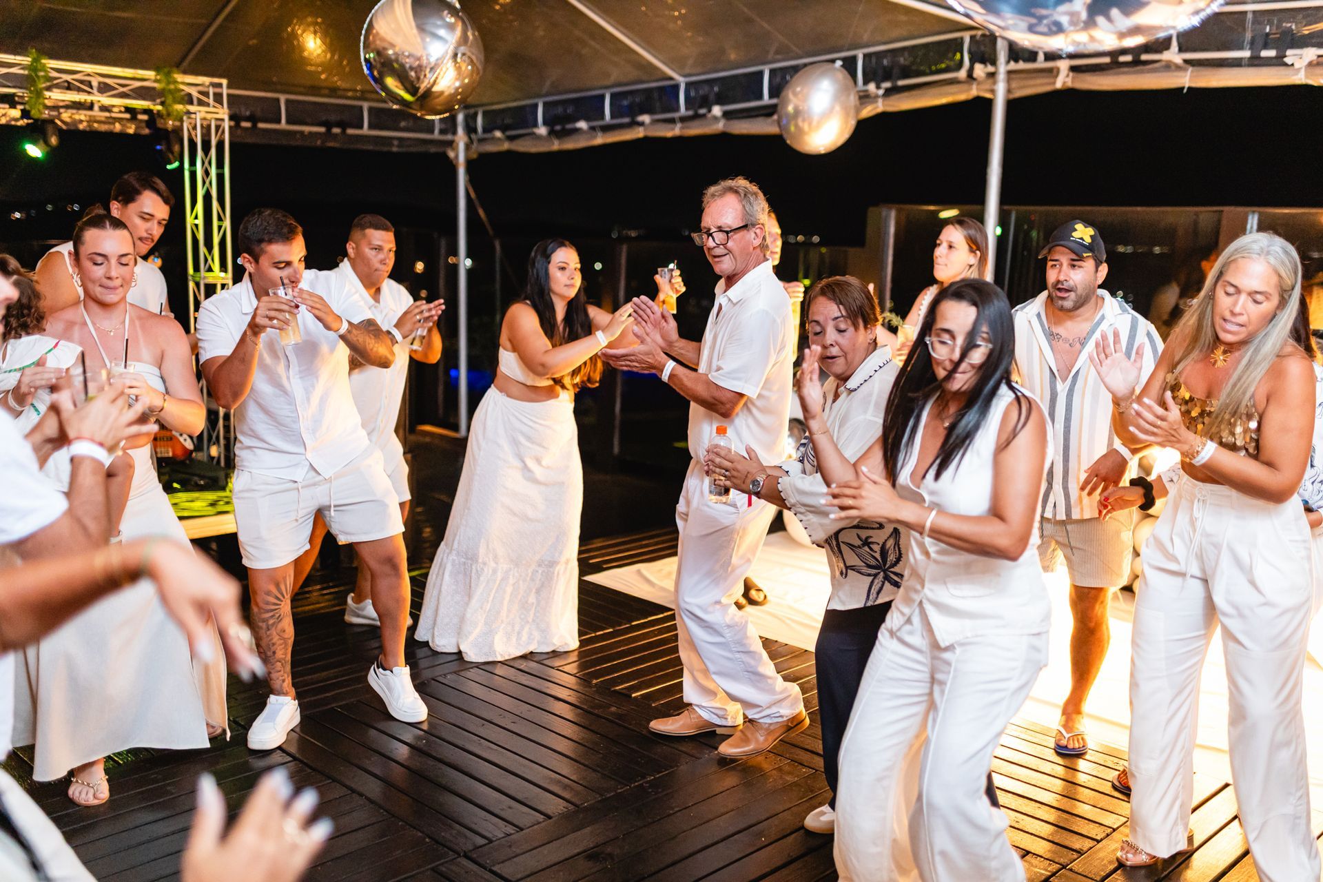 People dancing and clapping at outdoor party; many wear white clothing, disco balls hang.