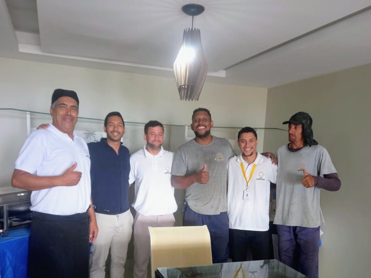 Grupo de seis pessoas sorrindo e fazendo sinal de positivo com o polegar em uma sala bem iluminada. Uma luminária está posicionada acima delas.