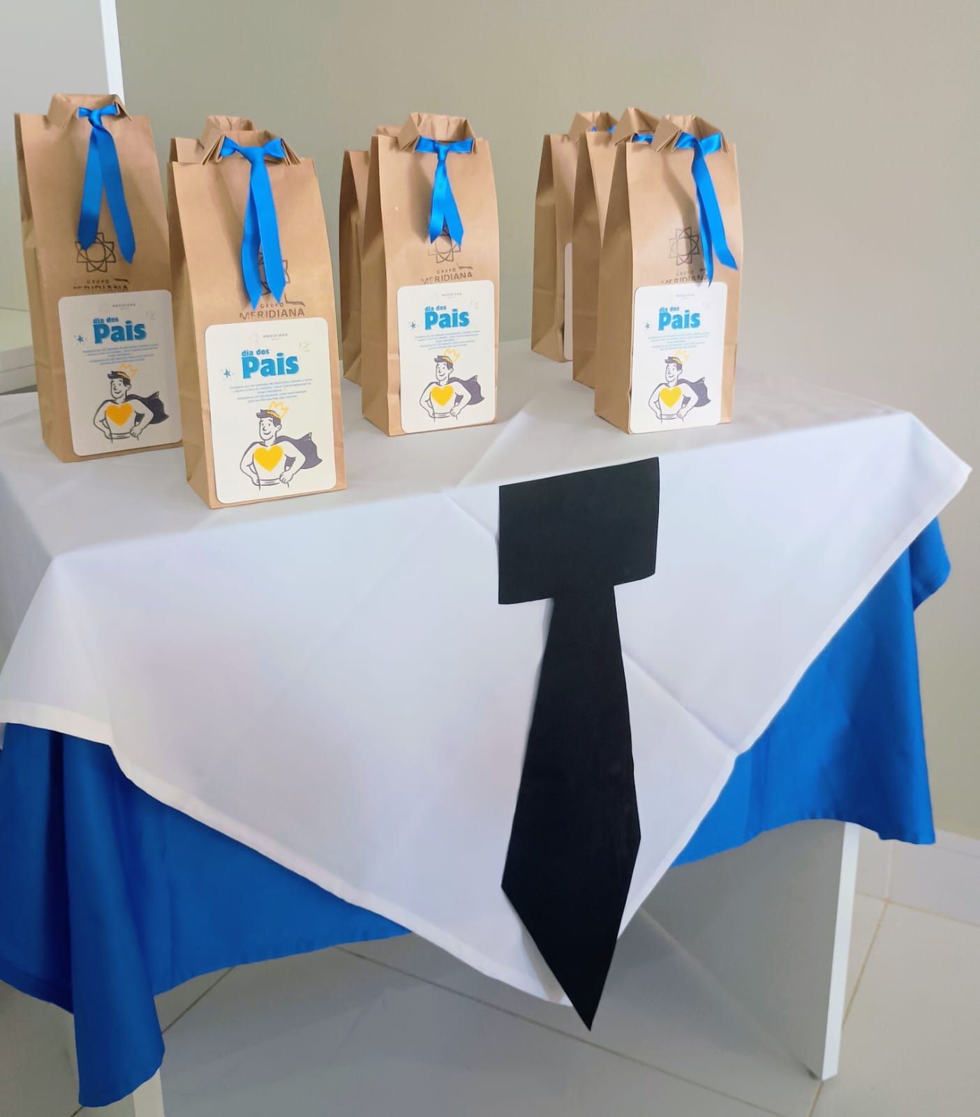Sacos de papel pardo amarrados com fitas azuis, com a etiqueta "Pais", sobre uma mesa com toalha de mesa branca e um desenho de gravata preta.