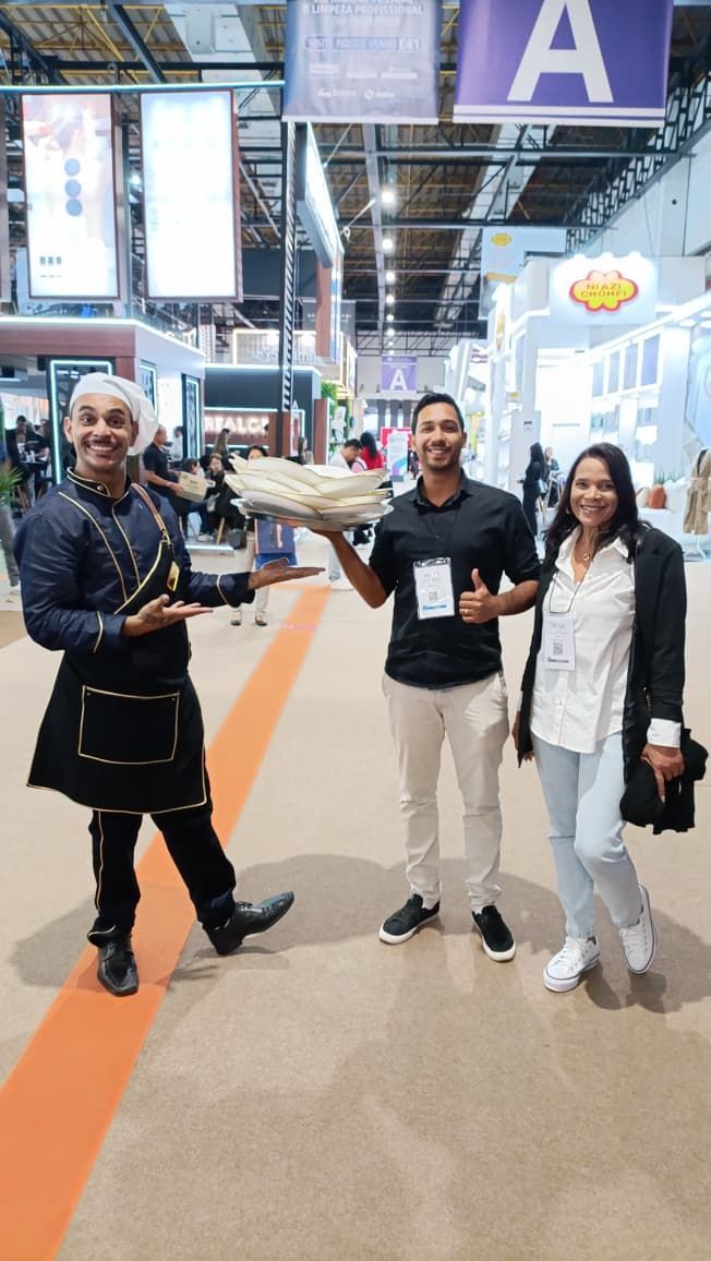 Um homem vestido de chef segura pão e posa para foto com duas pessoas em um centro de convenções.