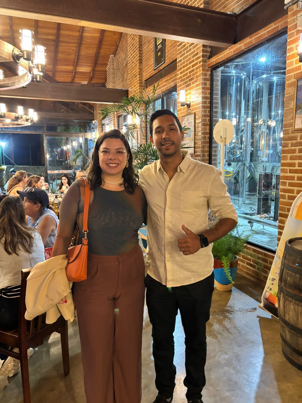 Duas pessoas sorrindo dentro de um restaurante. Mulher de calça marrom e homem de camisa social.