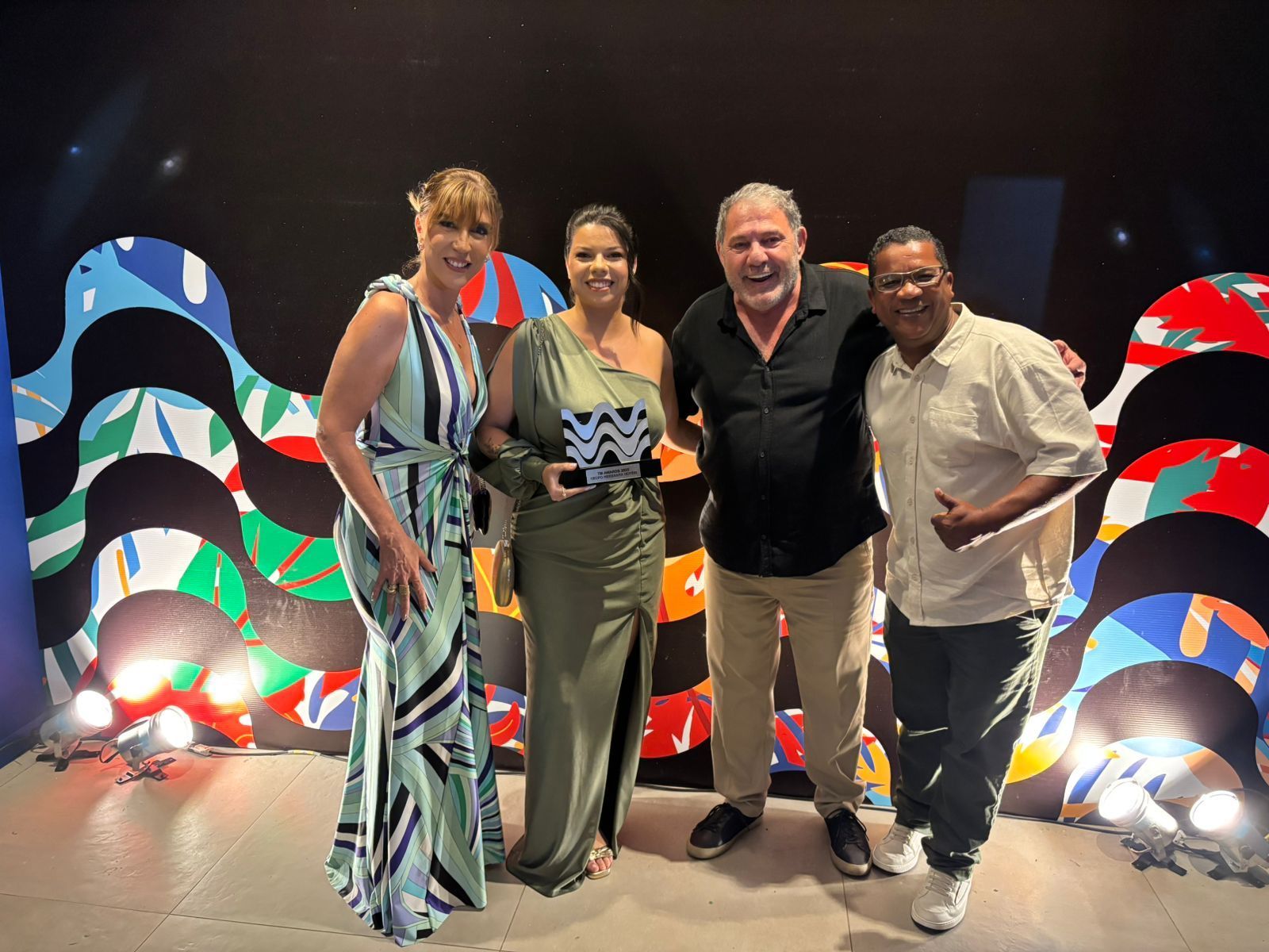 Quatro pessoas segurando um prêmio, sorrindo e posando em frente a um cenário colorido.