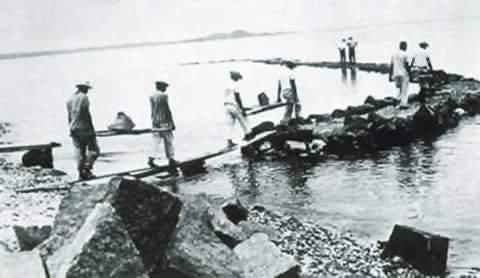 Homens trabalhando em um caminho de pedra na água, possivelmente uma ponte ou quebra-mar.