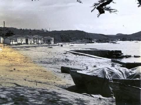 Cena de praia com pequenas construções, barcos e colinas ao fundo.