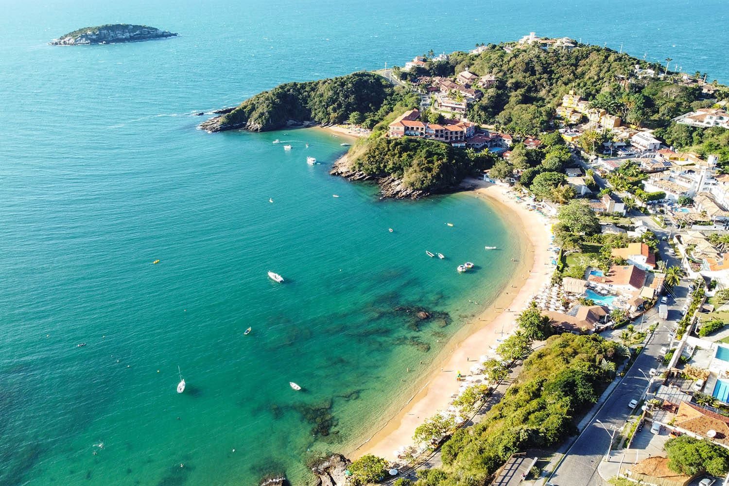 Aerial view of a coastal town with a sandy beach meeting turquoise water, surrounded by hills and scattered boats.