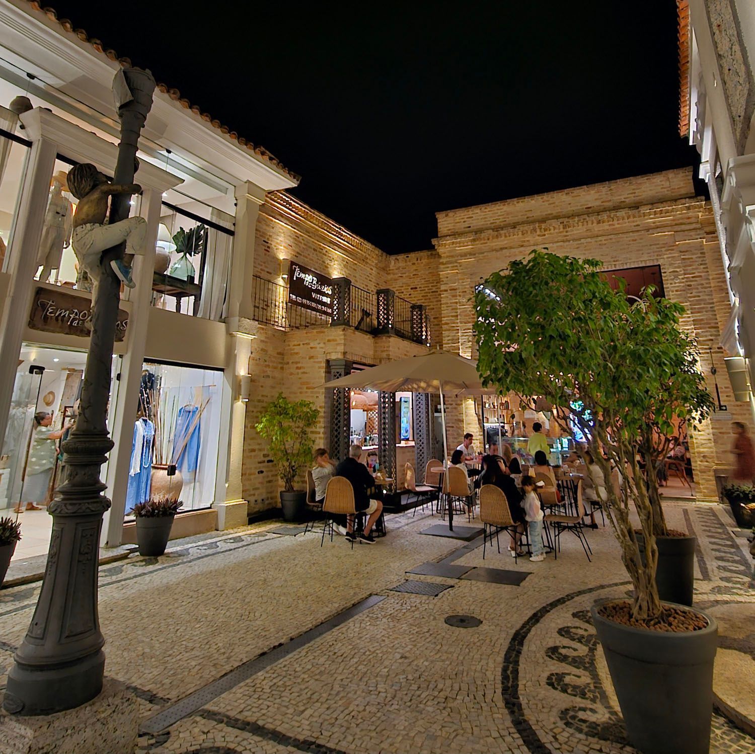 Vista noturna de uma área comercial com calçamento de pedra, vitrines iluminadas, um café e um poste de luz decorativo.