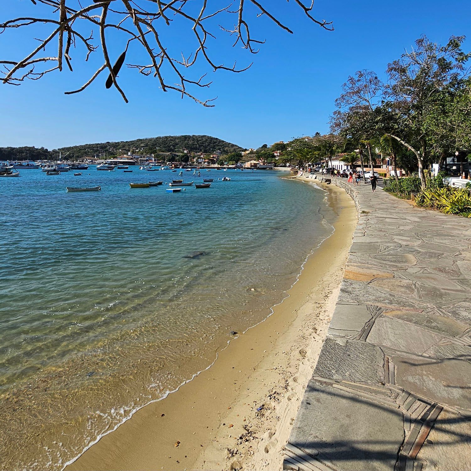 Praia de areia com barcos em águas calmas, caminho de pedra ao longo da costa, árvores à direita, colinas ao fundo.